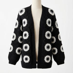 May include: Black open-front cardigan with a sunflower design. The sweater features a crochet pattern with white and gray flowers on a black background. The long-sleeved cardigan is displayed on a mannequin.