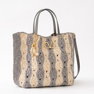 Pode incluir: Uma bolsa tote de crochê com listras verticais de fio creme e cinza. A bolsa possui duas alças de couro cinza, uma alça de ombro removível e um emblema com o logotipo V em tom dourado na frente. Um pingente dourado com borlas e pérolas pende de uma alça.