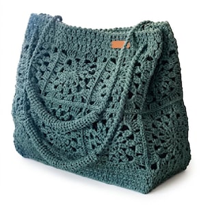 May include: A dark sage green tote bag constructed from crocheted granny squares. The bag features two matching crocheted shoulder straps and a small, rectangular, tan-coloured leather-like brand tag attached to the upper right front corner.
