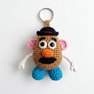 May include: A crochet keychain featuring a tan potato-shaped body with a black hat, pink ears, white arms, and blue feet. The figure has two black and white eyes, an orange nose, and a black mustache, all attached to a silver metal keyring.