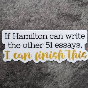 Op de afbeelding: Een witte sticker met zwarte tekst die luidt: "If Hamilton can write the other 51 essays, I can finish this." De woorden "I can finish this" zijn in gouden glitterletters.