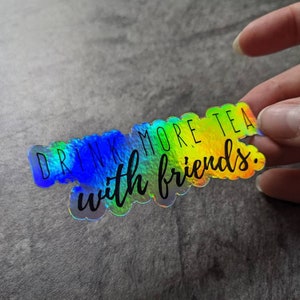 May include: Holographic sticker with the text "DRINK MORE TEA with friends." The sticker has a rainbow color effect, with blue, green, and yellow hues. The text is in a black, handwritten-style font.