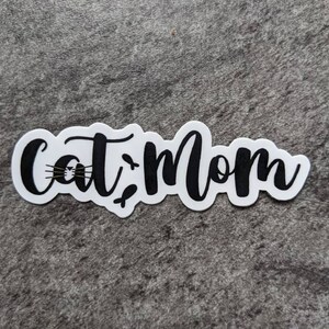 以下が含まれることがあります： 黒い筆記体で「Cat Mom」と書かれた白いステッカー。 「Cat」の文字にはヒゲのある猫の顔が描かれています。 ステッカーはグレーのテクスチャ加工された表面にあります。