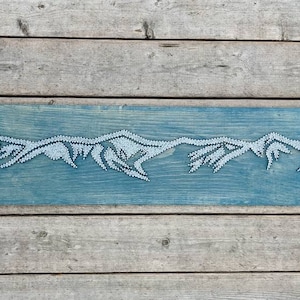May include: A rectangular wooden wall art piece featuring a mountain range design. The mountains are created with white string or thread against a teal blue painted background. The artwork is mounted on a wooden plank.