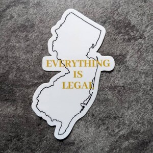 May include: White sticker in the shape of New Jersey with the text "EVERYTHING IS LEGAL" in gold lettering. The sticker has a black outline and is set against a dark gray, textured background.