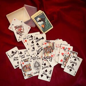 May include: A deck of playing cards with a clear plastic case and a card with a puffin on it. The cards are spread out on a red surface. Some cards have handwritten text.