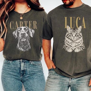 Op de afbeelding: Twee antracietgrijze T-shirts met korte mouwen. Het linker T-shirt toont een zwart-witte lijntekening van een hond met de naam CARTER erboven. Het rechter T-shirt toont een zwart-witte lijntekening van een kat met de naam LUCA erboven.