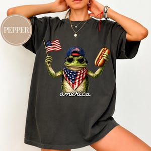 May include: A charcoal grey t-shirt featuring a graphic of a green frog wearing a blue baseball cap, heart-shaped sunglasses, and a red, white, and blue bandana. The frog holds a small American flag and a hot dog, with the text "amehica" below.