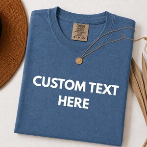 May include: A blue t-shirt with the text "CUSTOM TEXT HERE" in white. The shirt has a crew neck and a tag that says "COMFORT COLORS". A gold necklace with a pendant is draped across the shirt. A brown hat is visible in the upper left corner.