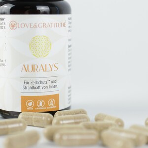 Pode incluir: Uma garrafa de cápsulas AURALYS com as palavras "Love & Gratitude" no rótulo. As cápsulas são bege e estão espalhadas ao redor da garrafa. O rótulo também inclui o texto "Für Zellschutz und Strahlkraft von Innen."