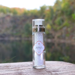 Könnte beinhalten: Eine klare Glasflasche mit Gesichtselixier, silberner Kappe und weißem Etikett. Auf dem Etikett steht "AURALYS Energetic For Youth Face Elixir" und ein Blumendesign. Die Flasche enthält 50 ml Produkt.