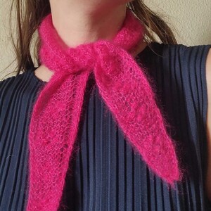 May include: A bright pink, fluffy scarf tied around the neck. The scarf is made of a soft, fuzzy material. The background is a dark blue, pleated top.