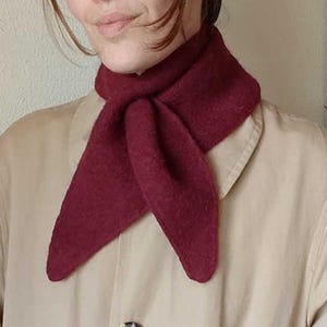 May include: A burgundy scarf is worn around the neck. The scarf is a deep red color and is knotted in the front. The person is wearing a tan coat with two visible buttons.