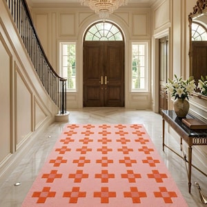 Puede incluir: Una alfombra rectangular con una base de color rosa claro y un patrón repetitivo de motivos en forma de cruz de color naranja dispuestos en cuadrícula. La alfombra está colocada sobre un suelo de mármol claro en un gran vestíbulo.