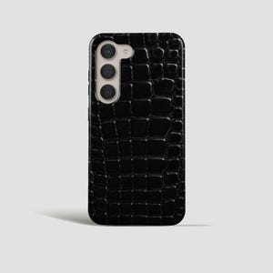 Peut inclure: Une coque de smartphone noire avec un motif texturé en peau de crocodile. La coque présente une forme rectangulaire aux coins arrondis et une découpe verticale sur le côté supérieur gauche pour loger un système de caméra à trois objectifs.