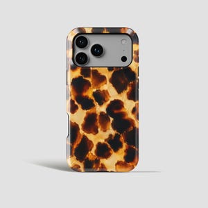 Op de afbeelding: Een smartphonehoesje met een glanzend schildpadpatroon met onregelmatige donkerbruine en amberkleurige vlekken. Het hoesje heeft een rechthoekige uitsparing linksboven voor een drievoudig camerasysteem en een flitser, met een lichtgrijs binnenframe.