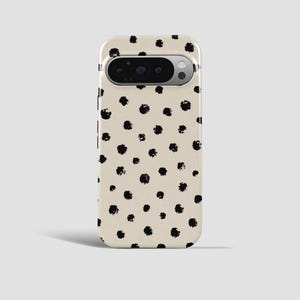 Op de afbeelding: Een smartphonehoesje met een crèmekleurige achtergrond en een patroon van onregelmatig geplaatste zwarte stippen. De hoes heeft een camera-uitsparing aan de bovenkant en is ontworpen voor een specifiek telefoonmodel.