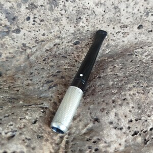 May include: A cigarette holder featuring a long, slender black stem and a textured silver-toned metal tip. The black portion includes a small, singular white dot detail near the transition point where it connects to the metallic end piece.