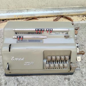 Może przedstawiać: Mechaniczny kalkulator vintage Everest Z5R w obudowie z metalu w przygaszonym oliwkowym kolorze. Urządzenie posiada klawiaturę numeryczną z klawiszami od 0 do 9 oraz dwa poziome okienka wyświetlacza z przesuwanymi czerwonymi i czarnymi wskaźnikami.