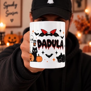 May include: A white ceramic mug featuring a Halloween-themed graphic with the text DADULA in a dripping red and black font. The design includes a ghost holding a coffee cup, black cats, orange pumpkins, bats, and red vampire-style wings.