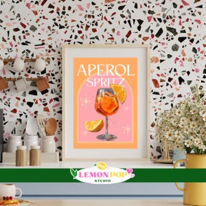 Op de afbeelding: Een ingelijste print met een illustratie van een Aperol Spritz cocktail. Het kunstwerk toont een feloranje cocktail in een wijnglas, gegarneerd met een sinaasappelschijfje, tegen een roze en oranje achtergrond. De tekst "APEROL SPRITZ" staat bovenaan.