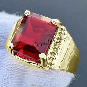 Könnte beinhalten: Ein Goldring mit einem großen, rechteckigen, facettierten roten Edelstein, der von vier Krappen gehalten wird. Die Ringschiene enthält eine Reihe kleiner, klarer, runder Steine an einer Seite. Der Text EliteGemsJewelry ist mittig zu sehen.