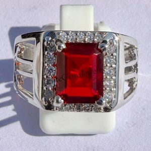 May include: A silver-toned ring featuring a large, rectangular, deep red center stone surrounded by a halo of small, clear, round-cut stones. The band has a wide, open-work design on the sides. The text EliteGemsJewelry is visible on the stone.