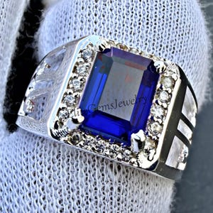 May include: A silver-toned ring featuring a large, rectangular, deep blue center stone surrounded by a halo of small, clear, round-cut stones. The band has a textured, open-work design on the sides. The text EliteGemsJewelry is visible.