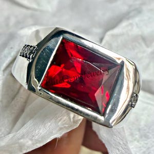 May include: A silver ring with a large, rectangular, faceted, red gemstone. The ring has a polished finish and a decorative design on the sides. The ring is displayed on a white surface. The text "Elite Gems Jewelry" is visible.