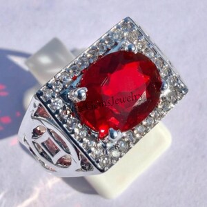 May include: A silver-toned ring featuring a large, oval-cut, vibrant red gemstone at the center. The rectangular setting is surrounded by a border of small, clear, sparkling stones. The band has a decorative cutout design and the text EliteGemsJewelry.