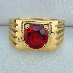 Pode incluir: Um anel de ouro com uma pedra preciosa vermelha escura, redonda e facetada, engastada numa moldura quadrada. A banda tem um acabamento dourado polido e largo com três ranhuras horizontais de cada lado. O texto EliteGemsJewelry é visível.