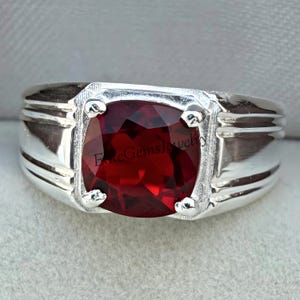 Puede incluir: Un anillo de plata con una gema roja oscura grande, de talla cuadrada. El anillo tiene una banda ancha con un acabado pulido y detalles decorativos alrededor de la gema. El texto "Elite Gems Jewelry" es visible.