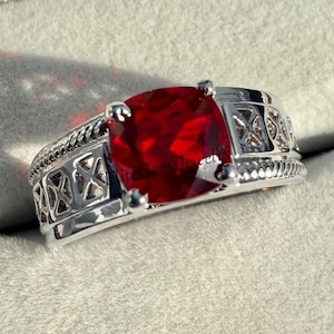 Könnte beinhalten: Ein silberfarbener Ring mit einem großen, runden, facettierten roten Edelstein, der von vier Krappen gehalten wird. Die Ringschiene zeigt ein detailliertes Gittermuster an den Seiten, eingefasst von einer strukturierten Kordeloptik an den Rändern.