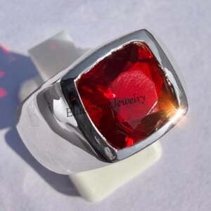 May include: A silver ring featuring a large, square-cut, faceted red gemstone set in a polished metal band. The surface of the red stone displays the text EliteGemsJewelry in black lettering. The ring has a smooth, wide, and reflective metal finish.