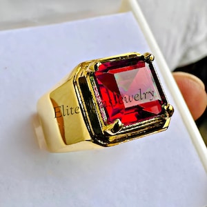 May include: A gold-toned ring featuring a large, square-cut, vibrant red gemstone held by four prongs. The band is smooth and polished. The text "Elite Gems Jewelry" is printed across the center of the red stone in a dark, thin font.