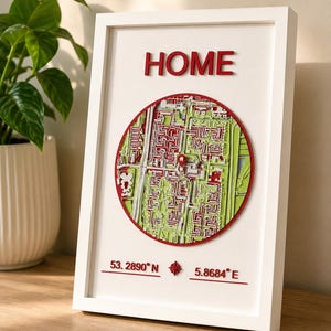 Op de afbeelding: Een wit ingelijst wandobject met een cirkelvormige 3D-kaart in rood- en groentinten. Het woord HOME staat in rode letters bovenaan. Onder de kaart staan de coördinaten 53.2890° N en 5.8684° E in rode tekst weergegeven.
