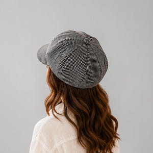 May include: A black and white houndstooth patterned newsboy cap with a rounded, multi-paneled crown and a small, stiff brim. The cap features a central fabric-covered button at the top and a structured shape that sits atop the head.