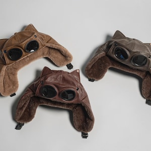 May include: Three aviator-style hats in shades of tan, brown, and dark brown. Each hat features cat-like ears on top, integrated dark-tinted goggles on the front, and a plush, fuzzy lining with ear flaps secured by black plastic buckles.