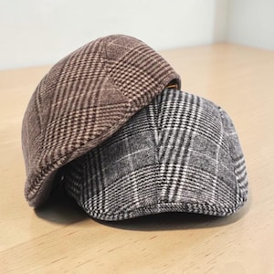 May include: Two flat caps stacked together. The top cap features a brown and tan plaid pattern, while the bottom cap displays a black, white, and gray plaid pattern. Both caps have a rounded crown and a short, stiff brim at the front.