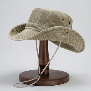 May include: A khaki-coloured wide-brimmed hat with a structured crown and a downward-sloping brim. The hat features metal ventilation eyelets on the sides, a brass-toned side snap button, and an adjustable chin strap with a black plastic toggle.
