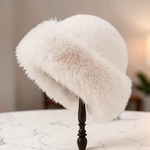 Może przedstawiać: Biały kapelusz typu bucket hat ze sztucznego futra z grubym, pluszowym rondem i zaokrągloną główką. Kapelusz ma miękką, gęstą fakturę i jednolity biały kolor. Konstrukcja obejmuje szerokie, wywinięte rondo, które otacza podstawę nakrycia głowy.