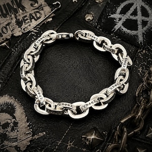 Puede incluir: Una pulsera de cadena plateada con texto grabado en cada eslabón. La pulsera está sobre una superficie de cuero negro con parches de temática punk y un imperdible. El texto "PUNK'S NOT DEAD" es visible.