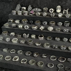 May include: A collection of silver rings displayed on tiered shelves. The rings feature various designs, including crosses, floral patterns, and geometric shapes. Some rings have gemstones. The rings are arranged on a dark gray surface.