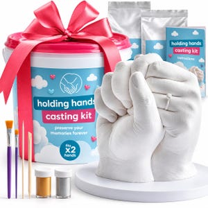 May include: A holding hands casting kit featuring a white bucket with a pink ribbon, two paint brushes, two wooden tools, two small paint jars, and a white plaster sculpture of two hands clasped together with a ring on one finger.