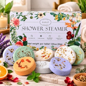 May include: A rectangular box labeled BLRIET SHOWER STEAMER with floral illustrations, containing multiple round, colorful shower steamer tablets embedded with dried flower petals, each weighing 1.05oz as stated on the packaging.