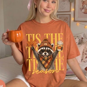 Könnte beinhalten: Ein orangefarbenes T-Shirt mit einer Grafik eines Waschbären, der eine Mütze, eine Sonnenbrille und einen karierten Schal trägt. Der Waschbär hält ein Ahornblatt und einen Kaffeebecher. Der Text 'TIS THE SEASON' steht in großen gelben Buchstaben.
