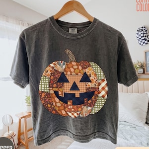 May include: A charcoal gray short-sleeve t-shirt featuring a large jack-o'-lantern graphic on the front. The pumpkin design is composed of various patchwork patterns in shades of orange, brown, green, and cream, with a dark navy blue face.