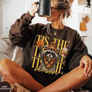 Könnte beinhalten: Ein dunkel schokoladenbraunes Langarm-Sweatshirt mit einer Grafik eines Waschbären, der eine Mütze, einen Schal und eine Sonnenbrille trägt. Der Waschbär hält ein Ahornblatt und einen Kaffeebecher. Der Text lautet TIS THE SEASON in Gelb.
