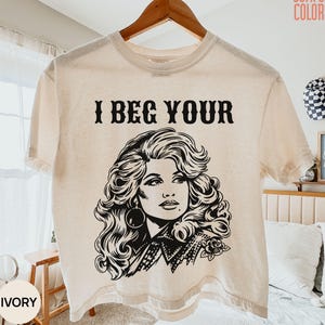 Könnte beinhalten: Ein elfenbeinfarbenes Kurzarm-T-Shirt mit einem schwarzen Grafikdruck einer Frau mit voluminösem Haar und dem Text I BEG YOUR über der Illustration. Das T-Shirt hat einen Rundhalsausschnitt und hängt an einem Holzkleiderbügel.
