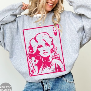May include: A light gray crewneck sweatshirt featuring a vibrant hot pink graphic print of a playing card design. The graphic depicts a stylized portrait of a woman with voluminous hair, the letter Q, a heart symbol, and a signature at the bottom.